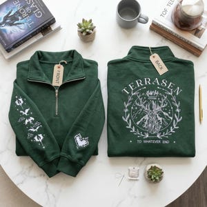May include: Two dark green sweatshirts with a quarter-zip collar are displayed. One has a front tag and white embroidered designs on the sleeve and chest. The other has a back tag and the word "TERRASEN" with a deer design.