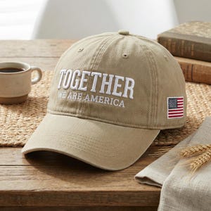 May include: A beige baseball cap with the embroidered text "TOGETHER WE ARE AMERICA" in white. The cap has a small American flag patch. The hat is displayed on a wooden surface.
