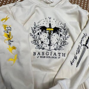 May include: Cream-colored hoodie with embroidered designs. Features a central emblem with a sword, wings, and celestial elements. The left sleeve has "Fly or die" with gold bird designs, and the right sleeve says "Violent Little Thing".