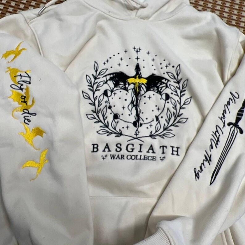 May include: Cream-colored hoodie with embroidered designs. Features a central emblem with a sword, wings, and celestial elements. The left sleeve has "Fly or die" with gold bird designs, and the right sleeve says "Violent Little Thing".