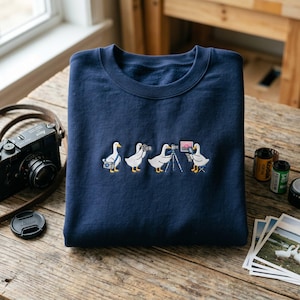 May include: Navy blue sweatshirt with an embroidered design of four ducks, each holding a different type of camera. The ducks are depicted in various stages of photography. The sweatshirt is folded on a wooden surface.