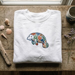 May include: A white crewneck sweatshirt featuring a central embroidered design of a manatee. The manatee's body is filled with a colorful pattern of coral, seaweed, and seashells in shades of orange, blue, green, and tan against the white fabric.