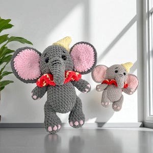 Puede incluir: Dos elefantes de ganchillo grises con grandes orejas forradas de rosa y detalles rojos y amarillos. Uno es más grande que el otro. Los elefantes tienen sombreros amarillos y bufandas rojas. Los peluches se exhiben contra una pared blanca.