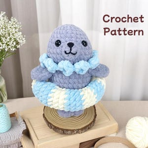 Puede incluir: Un peluche de foca de ganchillo en tonos grises, azules y blancos, con un flotador a rayas azules y blancas. La foca tiene ojos negros y una cara sonriente. El texto "Crochet Pattern" se muestra en marrón.