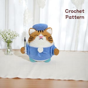 Puede incluir: Muñeco de gato tejido a ganchillo con uniforme y gorro de enfermera azules, sosteniendo una jeringa. El gato tiene pelaje naranja y blanco y una cara sonriente. El texto "Crochet Pattern" es visible.