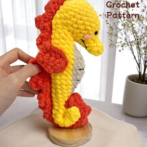 May include: A crocheted seahorse in shades of yellow, orange, and grey, displayed on a wooden base. The seahorse has a smiling face. The text "Crochet Pattern" is visible in the image. The seahorse is a handmade item.