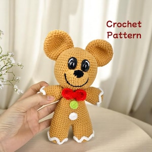 Może przedstawiać: Szydełkowy ludzik z piernika z uszami myszy, czerwoną muszką oraz zielonym i białym guzikiem. Piernikowy ludzik ma czarne oczy i uśmiechnięte usta. Tekst "Crochet Pattern" znajduje się w prawym górnym rogu.