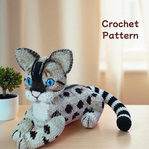 Peut inclure: Un chat en peluche crocheté avec des yeux bleus, des taches noires et une queue rayée. Le chat est couché et le texte "Crochet Pattern" est visible en arrière-plan.