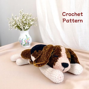 May include: A crocheted beagle dog plushie in brown, white, and black, lying down. The dog has long, floppy ears and a black nose. The text "Crochet Pattern" is visible in the image.