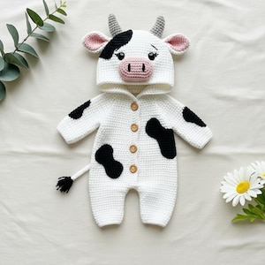 May include: A white crochet baby outfit designed to resemble a cow. The outfit features black spots, a hood with a cow face, and a small tail. The outfit has wooden buttons down the front.