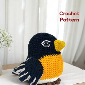 May include: A crocheted bird with a black head and wings, a yellow chest, and a yellow beak. The bird has white accents on the wings and a pair of large, round eyes. The text "Crochet Pattern" is in red on the right side.