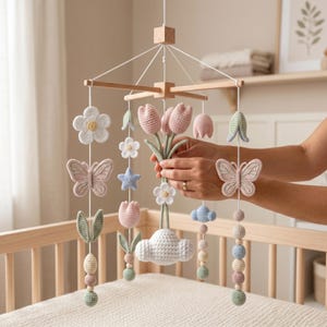 May include: A handmade baby mobile featuring crocheted flowers, butterflies, and clouds in pastel colors. The mobile includes pink tulips, white flowers, and a blue star, suspended from a wooden frame. The mobile is designed to hang over a crib.