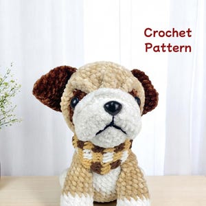 May include: A plush, crocheted dog with brown ears, a white face, and a tan body. It wears a checkered scarf in shades of brown and tan. The text "Crochet Pattern" is displayed in red.