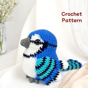 以下が含まれることがあります： 白、黒、ターコイズのアクセントが付いた、かぎ針編みの青い鳥。鳥は灰色のくちばしと足、大きな黒い目をしています。「Crochet Pattern」の文字が右上隅にあります。