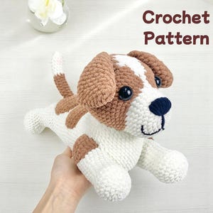 Puede incluir: Patrón de crochet para un amigurumi de perro marrón y blanco. El perro tiene ojos negros y una nariz negra. El perro está sentado en una mano.