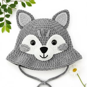May include: A gray crocheted wolf hat with white accents and black eyes. The hat features a wolf face design with a black nose and a white muzzle. The hat has ear flaps and a tie closure. The hat is made of a soft, textured yarn.