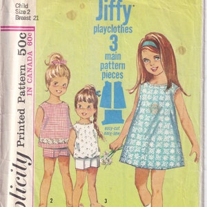Puede incluir: Un patrón de costura vintage Simplicity para ropa de juego para niños. El patrón es el número 6039 e incluye tres piezas principales. El patrón es para un niño de talla 2 con una medida de pecho de 21 pulgadas. El patrón está impreso en un fondo blanco con texto azul y verde. El patrón incluye una imagen de tres niñas usando la ropa de juego.