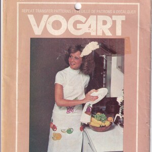 May include: A woman wearing a white apron with a fruit design is standing in a kitchen. She is holding a white plate in her left hand and a towel in her right hand. The towel has a purple eggplant design on it. There are grapes and bananas on the counter.