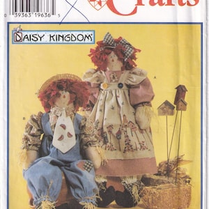 May include: Simplicity Crafts 7446 sewing pattern for two rag dolls. One doll wears blue overalls with a watermelon tie, and the other wears a tan dress with a birdhouse print. Both dolls have red yarn hair, straw accents, and black shoes.