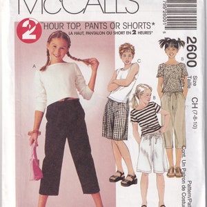 Op de afbeelding: McCall's naaipatroon-envelop met illustraties van kindertops, broeken en shorts. De envelop bevat de tekst "HOUR TOP, PANTS OR SHORTS" en maat informatie. De hoofdkleuren zijn wit, rood en zwart.