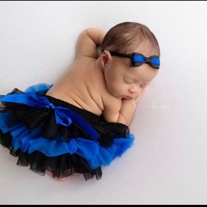 May include: A baby girl wearing a black and blue tutu with a blue bow headband. The baby is sleeping on a white background.