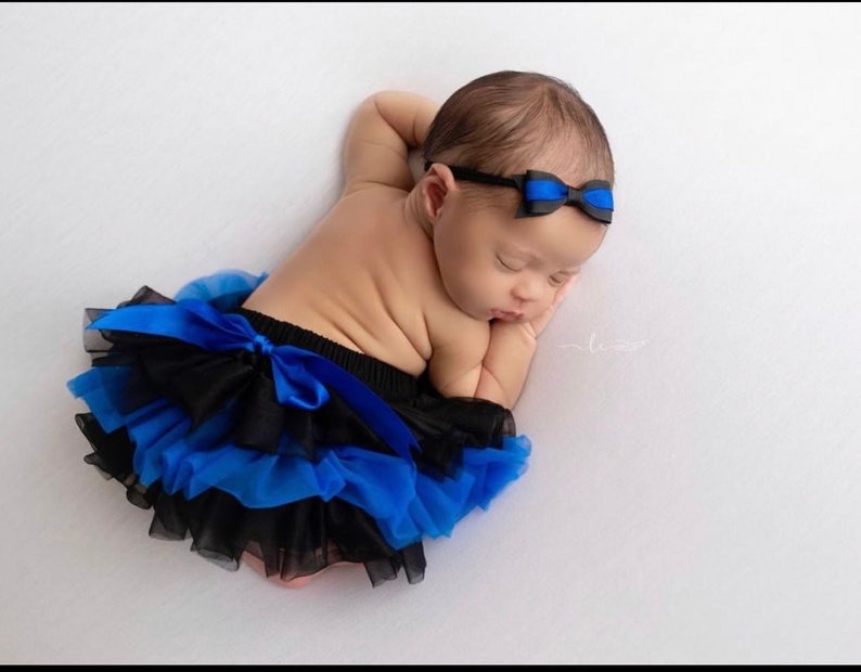 May include: A baby girl wearing a black and blue tutu with a blue bow headband. The baby is sleeping on a white background.