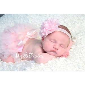 May include: A pink baby headband with a large pink flower on top. The headband is on a baby's head. The baby is wearing a pink tutu.