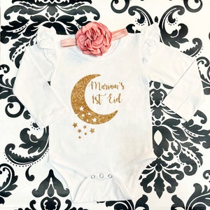 Op de afbeelding: Witte romper met lange mouwen met een gouden glitter halve maan en sterren. De romper heeft de tekst "Mariam's 1st Eid" erop.