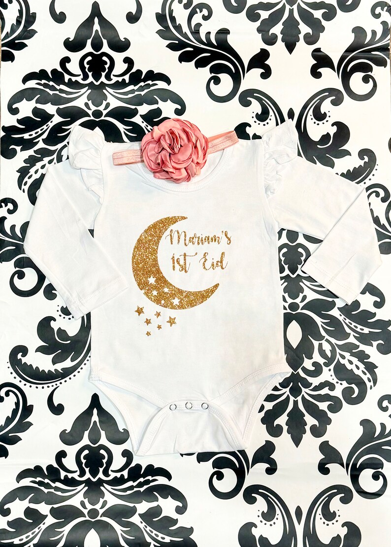 Op de afbeelding: Witte romper met lange mouwen met een gouden glitter halve maan en sterren. De romper heeft de tekst "Mariam's 1st Eid" erop.