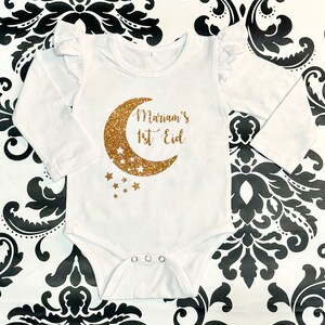 Op de afbeelding: Witte baby romper met lange mouwen met een gouden glitter halve maan en sterren. De romper heeft de tekst "Mariam's 1st Eid" erop.