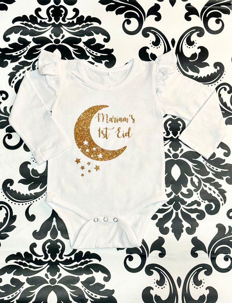 Op de afbeelding: Witte baby romper met lange mouwen met een gouden glitter halve maan en sterren. De romper heeft de tekst "Mariam's 1st Eid" erop.