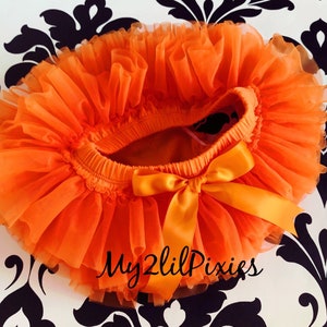 May include: Bright orange tutu skirt with a matching satin bow. The skirt is made of layers of tulle and has an elastic waistband. The text "My2lilPixies" is written in cursive on the bottom right of the skirt.