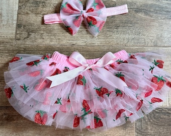 May include: Pink tulle tutu skirt with a pink bow and strawberry print. The skirt is layered and has a pink elastic waistband. A matching pink headband with a strawberry bow is included.