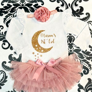 Op de afbeelding: Een witte romper met lange mouwen met een ontwerp van een gouden glitter halve maan en sterren. De romper heeft de tekst "Mariam's 1st Eid" erop. De romper wordt gecombineerd met een roze tule tutu.