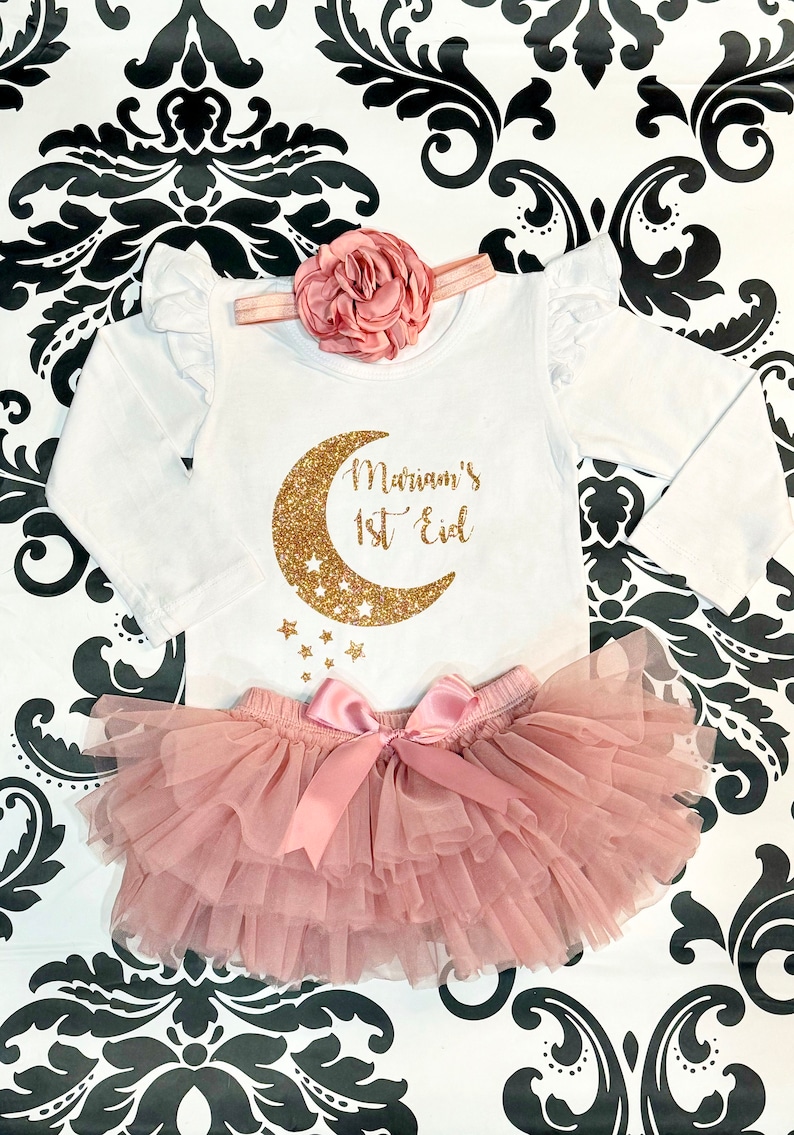 Op de afbeelding: Een witte romper met lange mouwen met een ontwerp van een gouden glitter halve maan en sterren. De romper heeft de tekst "Mariam's 1st Eid" erop. De romper wordt gecombineerd met een roze tule tutu.