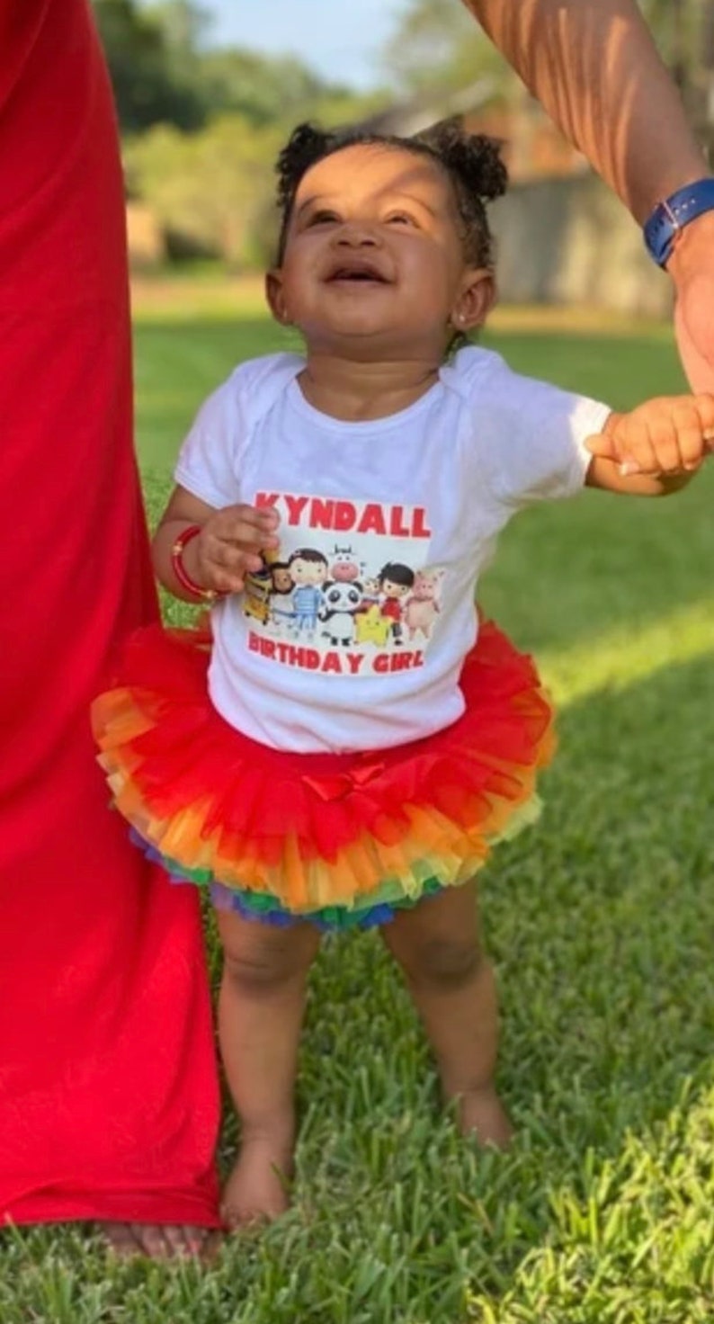 Puede incluir: Una ni&ntilde;a peque&ntilde;a con una camiseta blanca con el texto "Kyndall Birthday Girl" y una imagen de dibujos animados de personajes del programa "Cocomelon". La ni&ntilde;a tambi&eacute;n lleva una falda de tul roja y arco&iacute;ris.