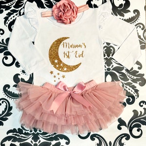 Op de afbeelding: Een witte romper met lange mouwen met een ontwerp van een gouden glitter halve maan en sterren. De romper zegt "Mariam's 1st Eid". Een roze tule tutu met een roze satijnen strik is inbegrepen.