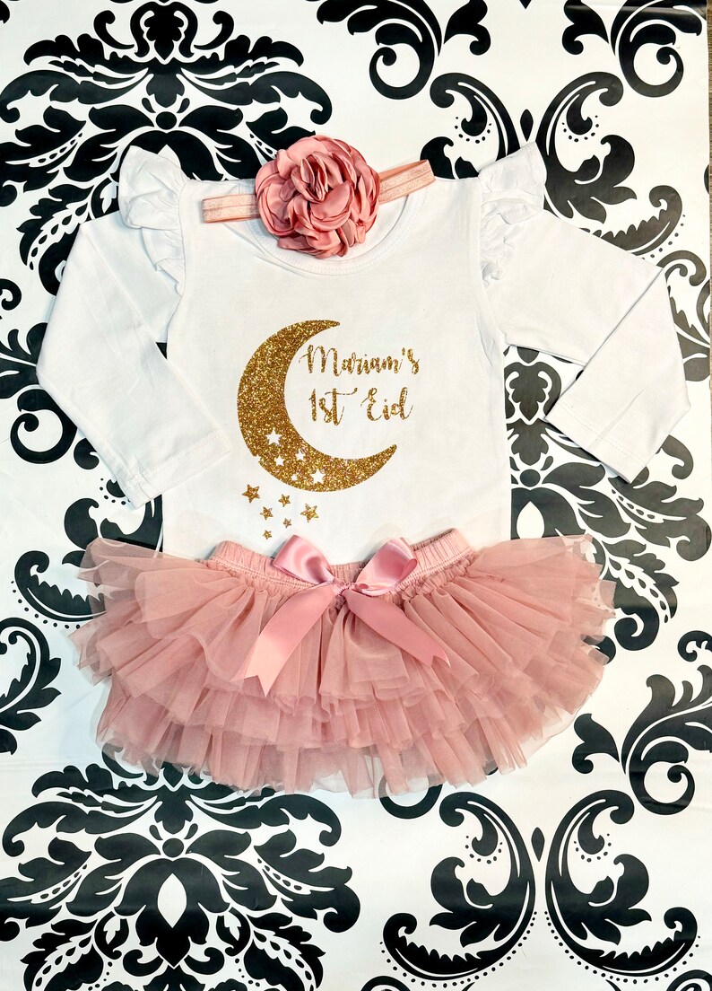 Op de afbeelding: Een witte romper met lange mouwen met een ontwerp van een gouden glitter halve maan en sterren. De romper zegt "Mariam's 1st Eid". Een roze tule tutu met een roze satijnen strik is inbegrepen.