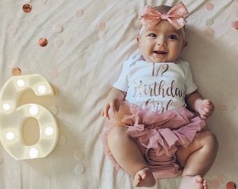 May include: A baby girl wearing a white onesie with the text "1/2 Birthday Girl" in rose gold lettering. She is wearing a pink tutu and a pink bow headband. The baby is lying on a white blanket with pink and gold confetti.