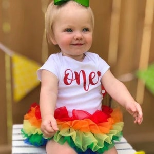 Puede incluir: Una ni&ntilde;a peque&ntilde;a con un body blanco con la palabra "one" en purpurina roja en la parte delantera. Tambi&eacute;n lleva una falda de tul arco&iacute;ris con tul rojo, naranja, amarillo, verde, azul y morado. Lleva un lazo verde en el pelo.