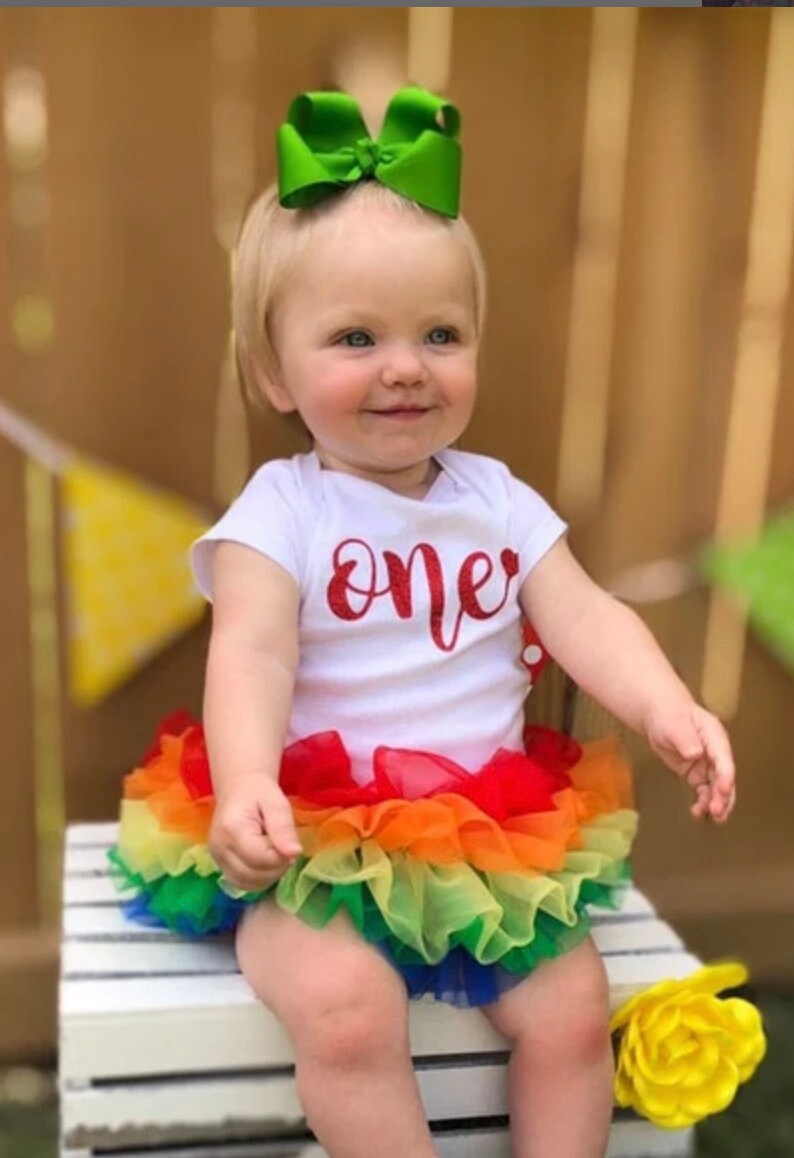 Puede incluir: Una ni&ntilde;a peque&ntilde;a con un body blanco con la palabra "one" en purpurina roja en la parte delantera. Tambi&eacute;n lleva una falda de tul arco&iacute;ris con tul rojo, naranja, amarillo, verde, azul y morado. Lleva un lazo verde en el pelo.