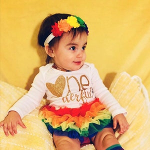 Puede incluir: Una ni&ntilde;a peque&ntilde;a con un vestido blanco de manga larga con letras doradas brillantes que dicen "One Derful" y un coraz&oacute;n. Tambi&eacute;n lleva una falda de tul de arco&iacute;ris y una diadema de flores de arco&iacute;ris.