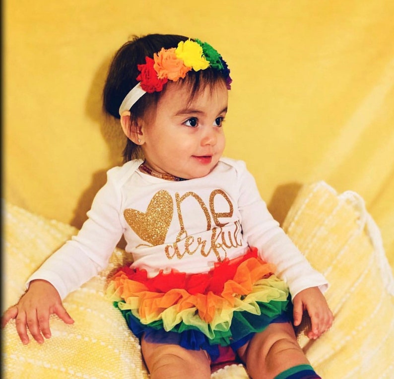 Puede incluir: Una ni&ntilde;a peque&ntilde;a con un vestido blanco de manga larga con letras doradas brillantes que dicen "One Derful" y un coraz&oacute;n. Tambi&eacute;n lleva una falda de tul de arco&iacute;ris y una diadema de flores de arco&iacute;ris.