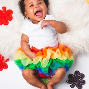 Puede incluir: Una ni&ntilde;a peque&ntilde;a con un body blanco y una falda de tul arco&iacute;ris. Est&aacute; riendo y sentada sobre una superficie blanca y esponjosa con formas de hojas de fieltro de colores alrededor.