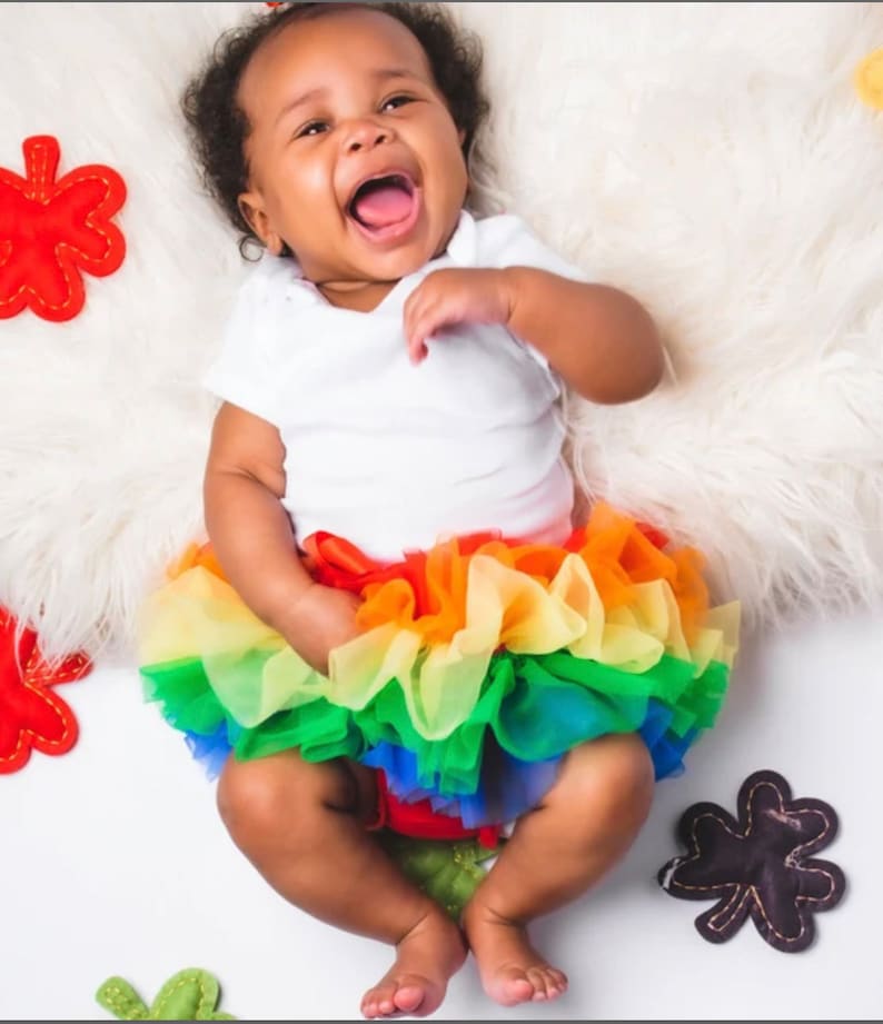 Puede incluir: Una ni&ntilde;a peque&ntilde;a con un body blanco y una falda de tul arco&iacute;ris. Est&aacute; riendo y sentada sobre una superficie blanca y esponjosa con formas de hojas de fieltro de colores alrededor.