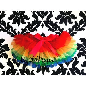 Puede incluir: Tutu de tul arco&iacute;ris con un lazo de sat&eacute;n rojo. El tutu tiene varias capas de tul en los colores del arco&iacute;ris, con una capa roja en la parte superior. El tutu est&aacute; sobre un fondo estampado en blanco y negro.