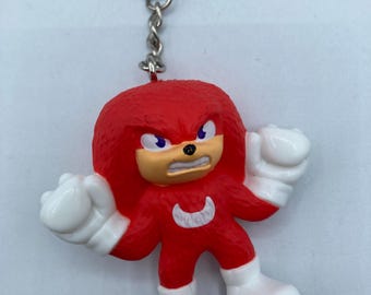 May include: A red Knuckles the Echidna keychain figurine with white gloves and boots. The figurine has a stern expression with purple eyes and a tan muzzle. The keychain includes a silver chain and ring.