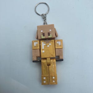 May include: A tan and gold keychain featuring a pixelated pig character. The figure has a square head with small ears and a snout. The body is gold with white accents. The keychain has a silver ring and chain.