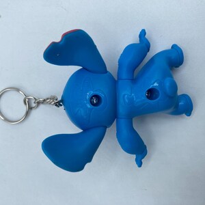 K&ouml;nnte beinhalten: Ein blauer Stitch-Schl&uuml;sselanh&auml;nger mit den charakteristischen Merkmalen der Figur, darunter gro&szlig;e Ohren, ein runder K&ouml;rper und vier Gliedma&szlig;en. Der Schl&uuml;sselanh&auml;nger hat einen silbernen Metallring und eine Kette. Die Ohren der Figur haben rote Akzente.