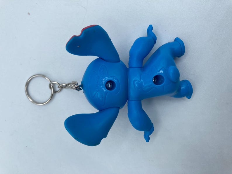 K&ouml;nnte beinhalten: Ein blauer Stitch-Schl&uuml;sselanh&auml;nger mit den charakteristischen Merkmalen der Figur, darunter gro&szlig;e Ohren, ein runder K&ouml;rper und vier Gliedma&szlig;en. Der Schl&uuml;sselanh&auml;nger hat einen silbernen Metallring und eine Kette. Die Ohren der Figur haben rote Akzente.